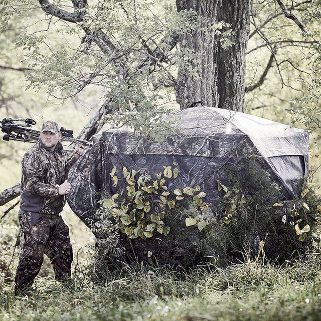 Ameristep Pro Series 56x56 Extreme View ground blind set up in wooded area with hunter in camouflage gear holding crossbow beside the blind