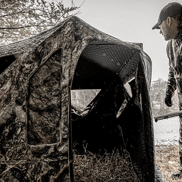 Ameristep Pro Series Insulated 59x59 pop-up ground blind with hunter in camouflage gear standing beside the open door in a wooded field