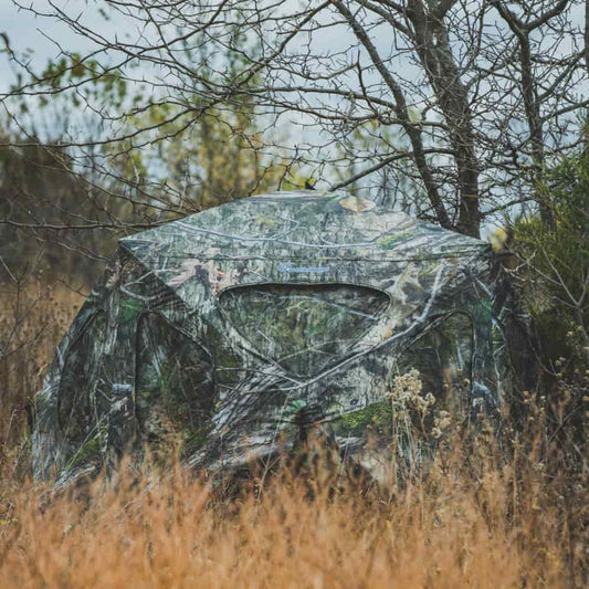 Ameristep Silent Brickhouse 59x59 pop-up ground blind in Mossy Oak camouflage set up in tall grass and wooded field