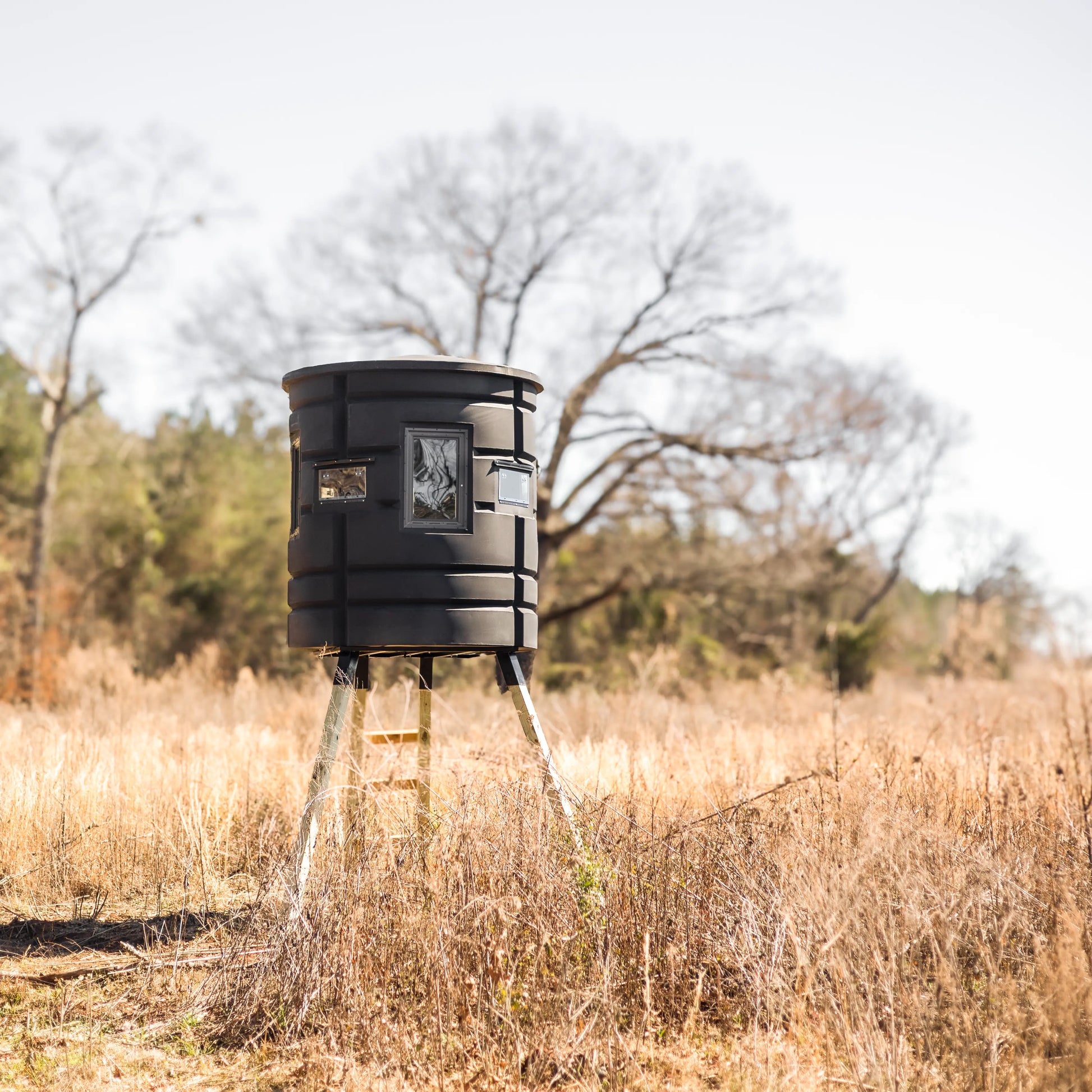 Wide shot of Sportsman Bow Condo elevated deer blind in an open grassy field with trees in the background