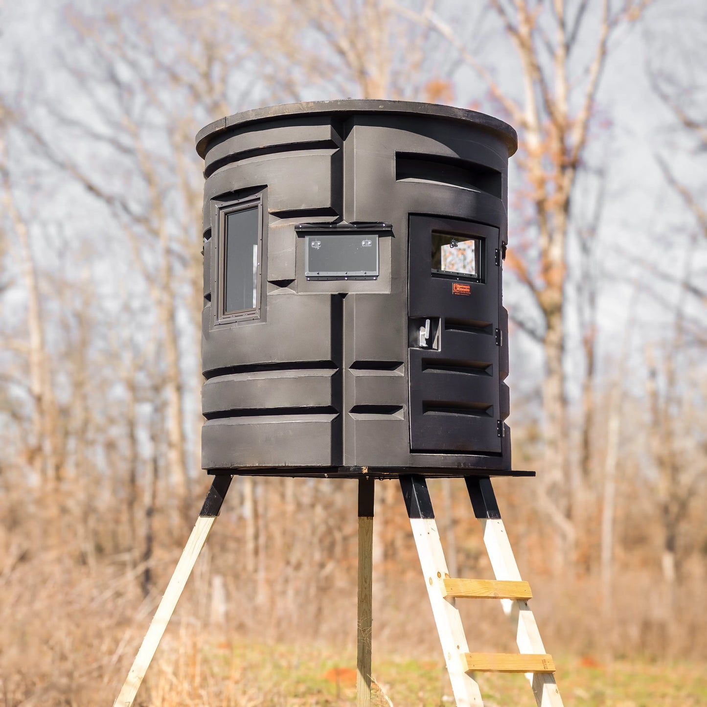 Front angle of Sportsmans Condo Bow Condo elevated deer blind with ladder and door visible in a wooded area