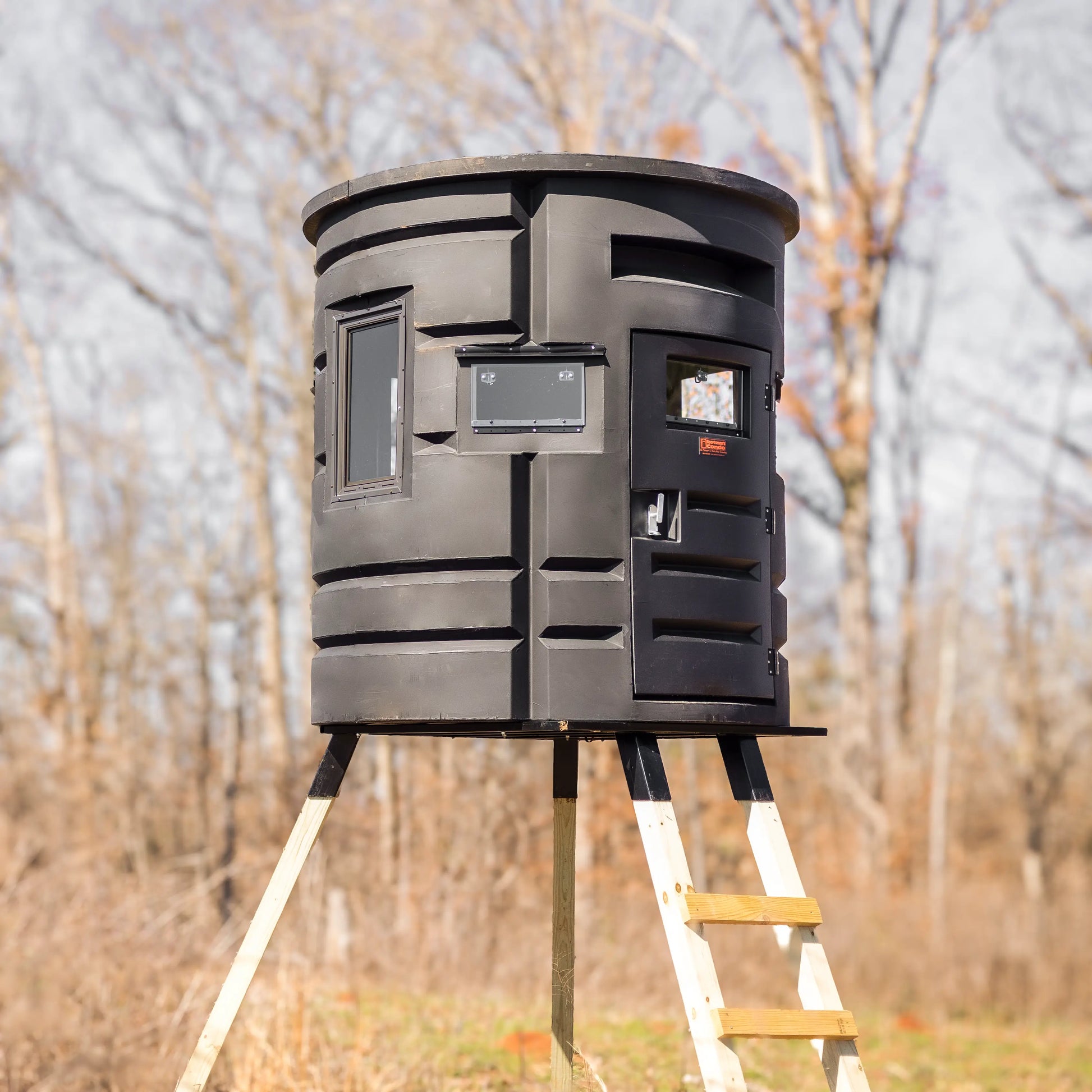Front angle of Sportsmans Condo Bow Condo elevated deer blind with ladder and door visible in a wooded area