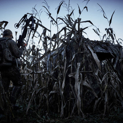 Ameristep Care Taker Run & Gun 49x49 pop-up ground blind set up in a cornfield with hunter in camouflage gear holding a compound bow at dusk