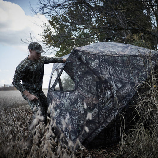 Man in camouflage clothing interacting with a camouflaged hunting blind in a field.