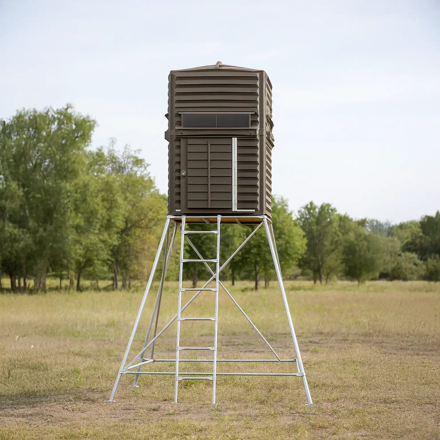 The Blynd 4x4 Deer Hunting Blind elevated on tall steel stand in an open grassy field, providing excellent visibility for rifle or crossbow hunting.

