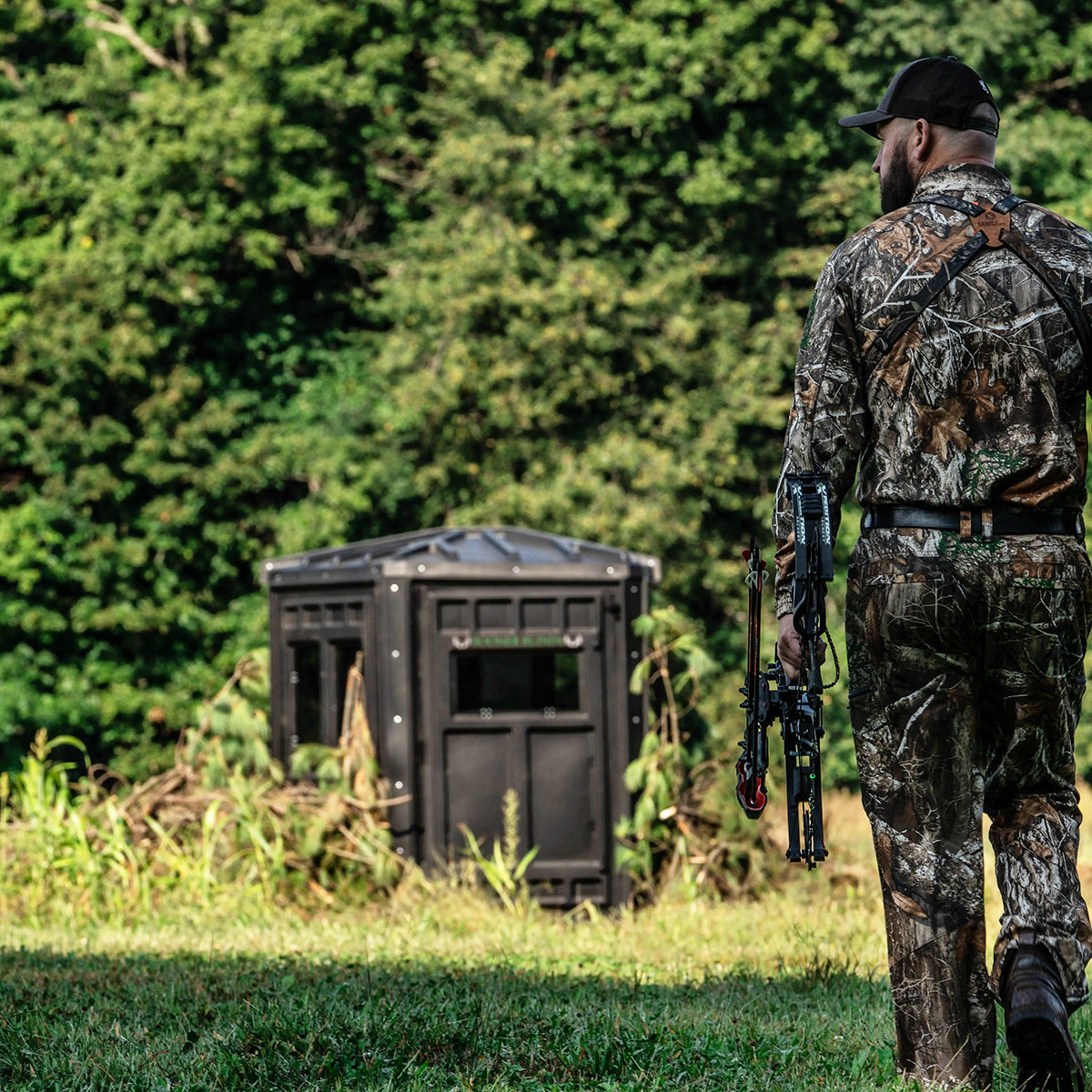 Bowhunter walking toward a Booner 6-Panel deer hunting blind in a field setting, ground blind setup for whitetail hunting sold by Outdoors For Less