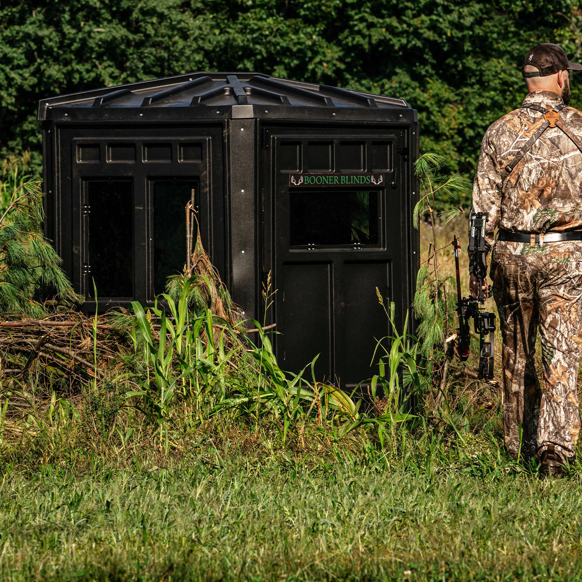 Bowhunter standing beside a Booner 6-Panel deer hunting blind brushed into natural cover, concealed ground blind setup sold by Outdoors For Less