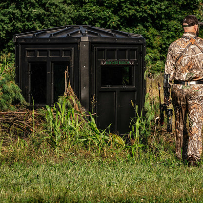 Bowhunter standing beside a Booner 6-Panel deer hunting blind brushed into natural cover, concealed ground blind setup sold by Outdoors For Less