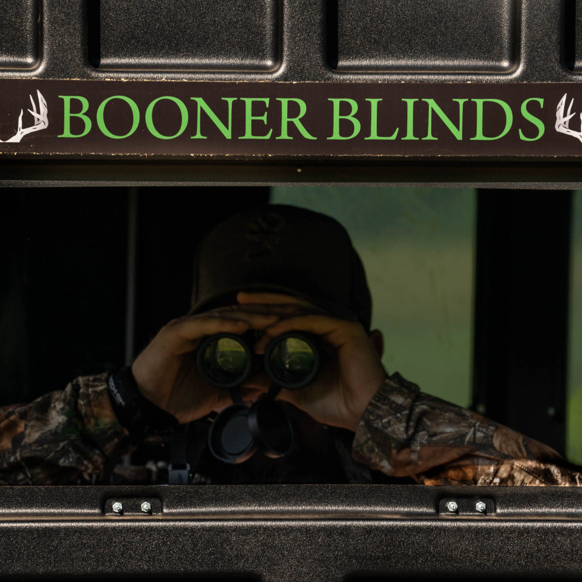 Hunter using binoculars inside a Booner deer hunting blind with Booner Blinds logo visible, premium whitetail hunting blind sold by Outdoors For Less