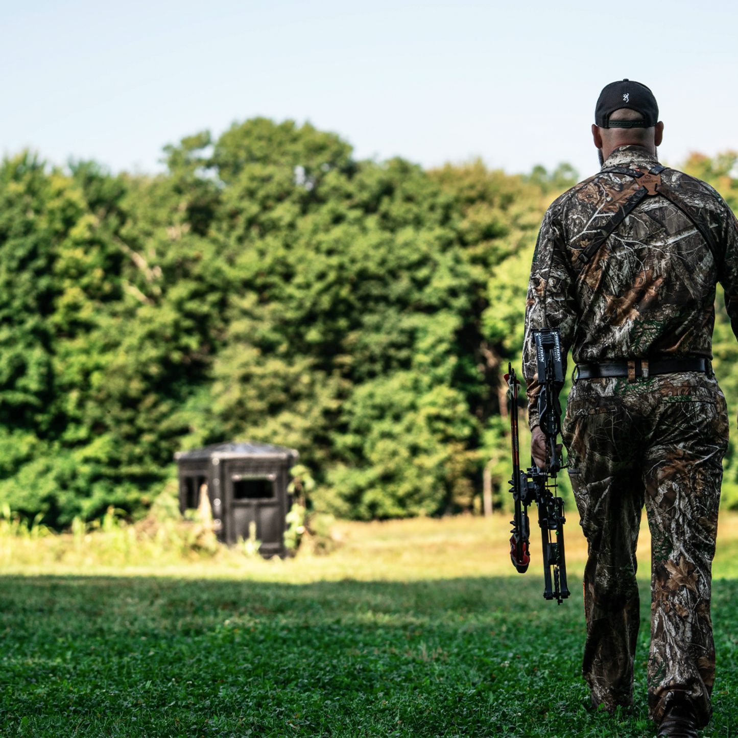Bowhunter walking toward a Booner deer hunting blind in a grassy field, ground blind setup for whitetail hunting sold by Outdoors For Less