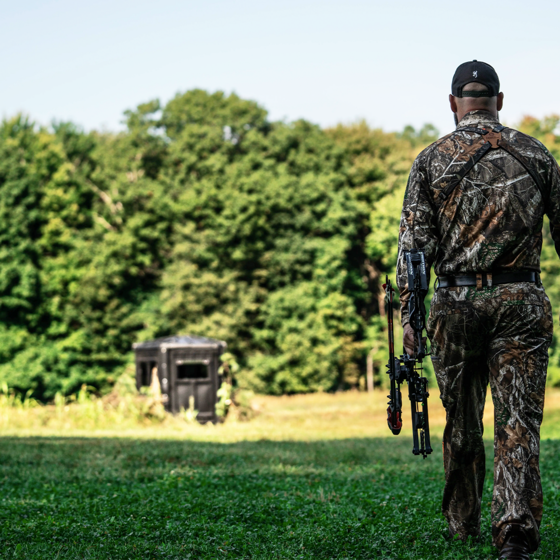 Bowhunter walking toward a Booner deer hunting blind in a grassy field, ground blind setup for whitetail hunting sold by Outdoors For Less