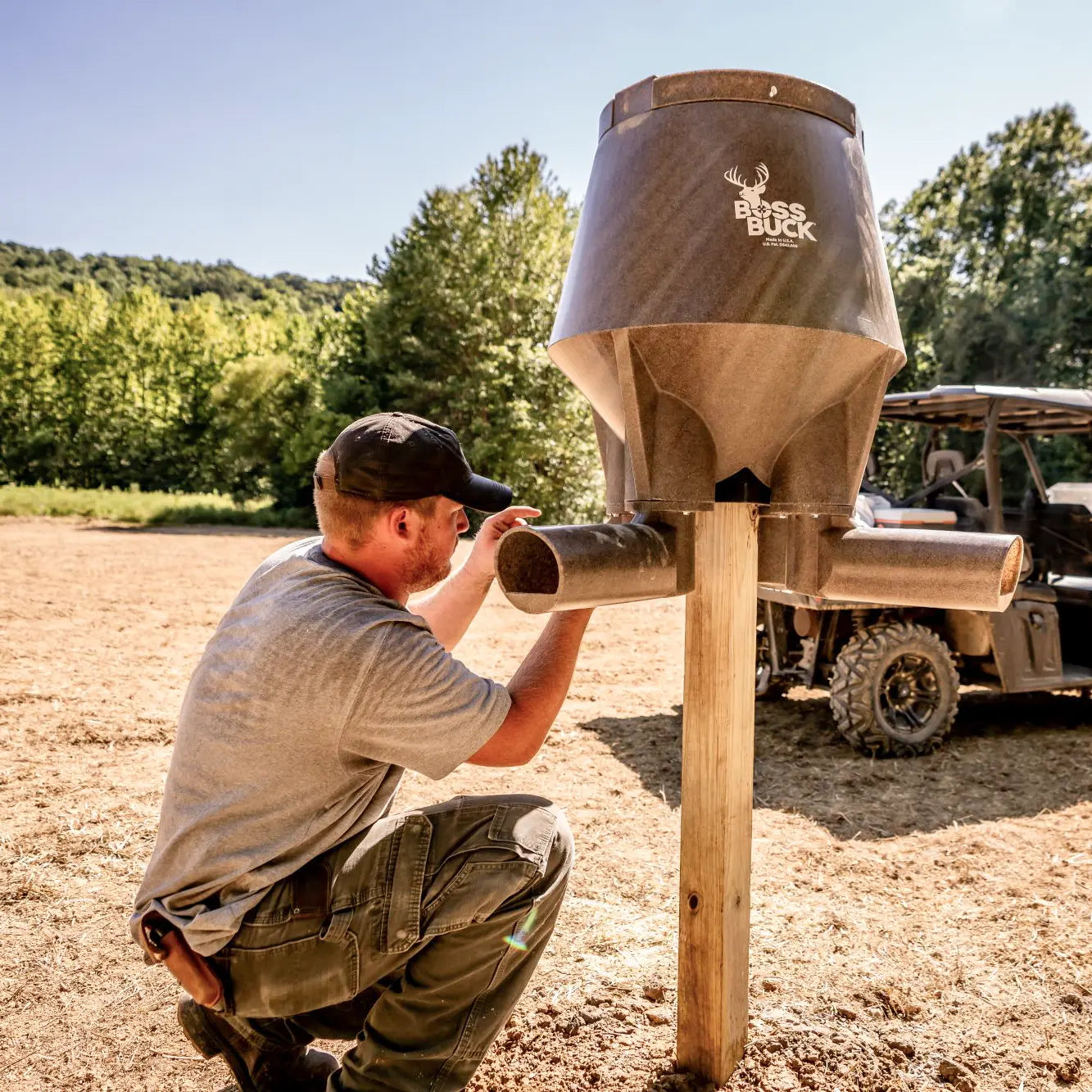 Boss Buck 200lb Post-Mounted Deer Feeder on 4x4 post with hunter kneeling beside — durable brown gravity feeder made from rotomolded plastic for long-lasting deer feeding setups.