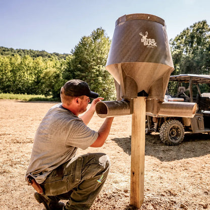 Boss Buck 200lb Post-Mounted Deer Feeder on 4x4 post with hunter standing beside — brown rotomolded gravity feeder for deer management and wildlife feeding.