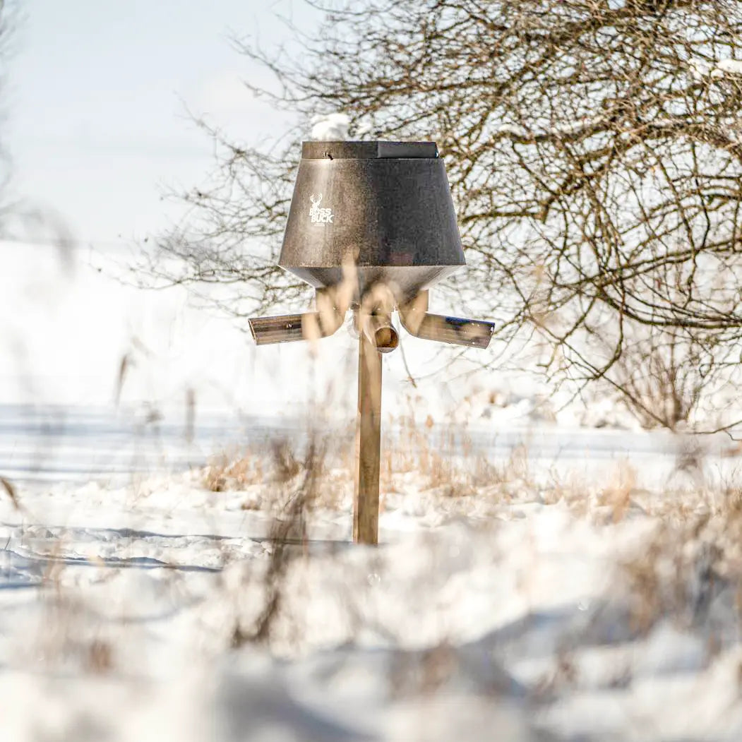 Boss Buck 200lb Post-Mounted Deer Feeder in snowy winter landscape — gravity feed design provides reliable wildlife feeding during all seasons, featuring UV-resistant brown plastic hopper.