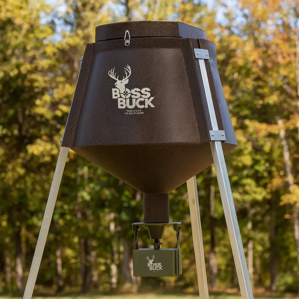 Boss Buck 350 lb Automatic Deer Feeder set up in forest clearing — sturdy brown rotomolded deer feeder with tripod legs, 12-volt digital timer, and automatic spinner plate.