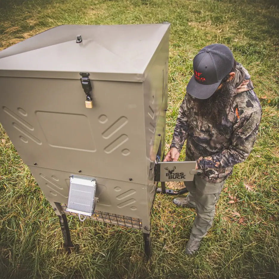 Hunter standing beside Boss Buck 600lb Stand & Fill Automatic Sled Deer Feeder — tan feeder with 12-volt spinner system, digital timer, and heavy-duty sled base for all-terrain stability.