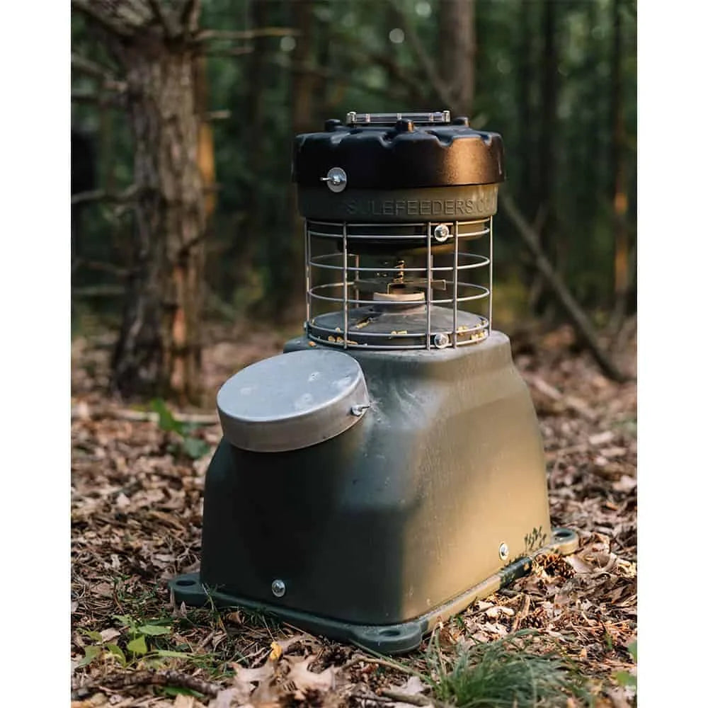 Capsule Feeders 100-LB Easy-Fill Game Feeder placed on forest floor among leaves and pine cones, side view