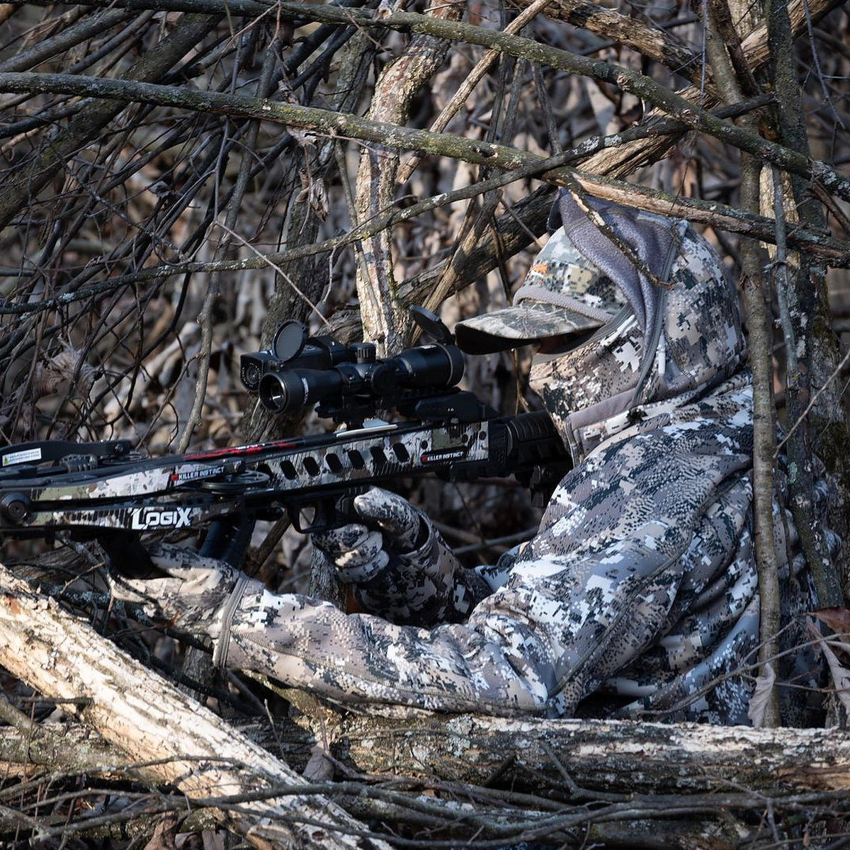 Hunter in camouflage aiming Killer Instinct crossbow while concealed in natural brush during hunting setup