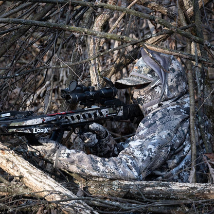 Hunter in camouflage aiming Killer Instinct crossbow while concealed in natural brush during hunting setup
